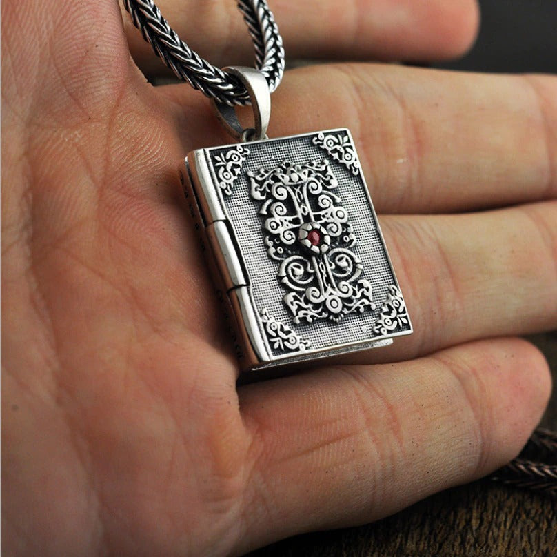 Vintage silver pendant shaped like a religious book, detailed with ornate designs and a red stone accent, showcased in hand.