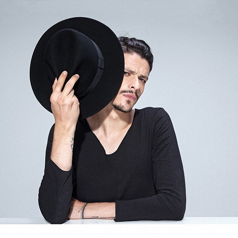 Man in black shirt playfully holding a wide brim wool fedora, showcasing alternative fashion style.
