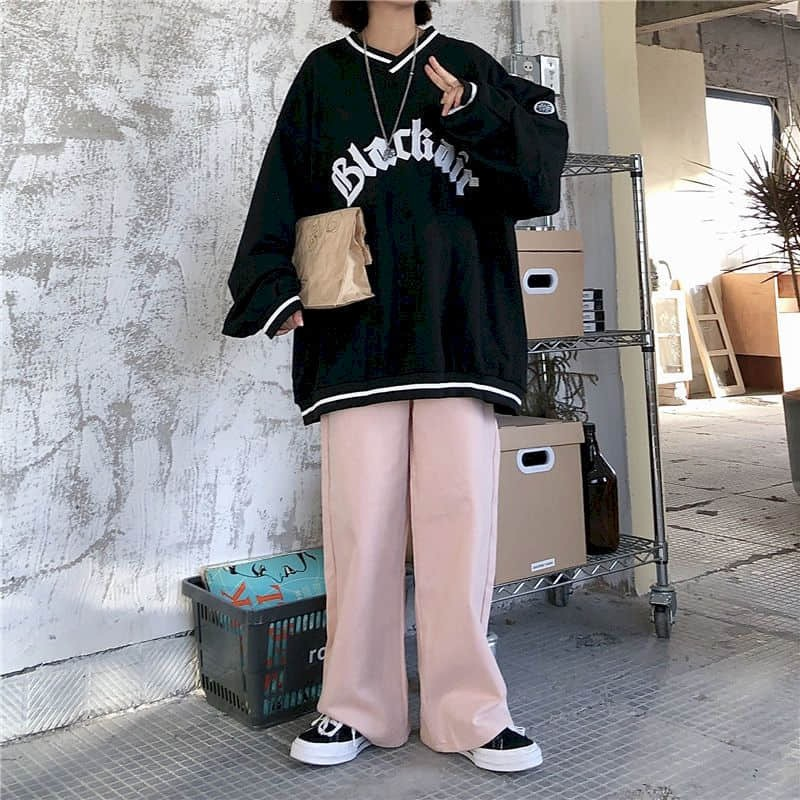 Fashionable woman in oversized V-neck black sweatshirt with letter patch, paired with pink pants and sneakers.