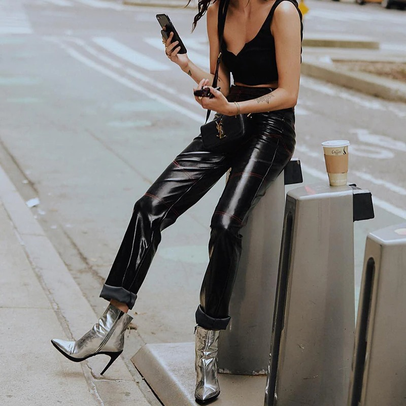 Trendy woman in black PU leather pants, silver boots, and crop top, checking phone while sitting on a city street.